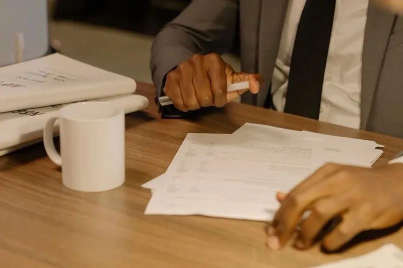 A person in a suit holds a pen over printed statement pages spread across a wooden table, a second person&rsquo;s hand resting on the paperwork