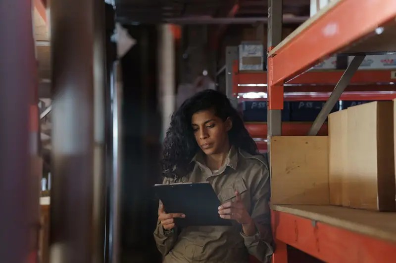 A worker checks a clipboard while counting stock in a dim warehouse aisle