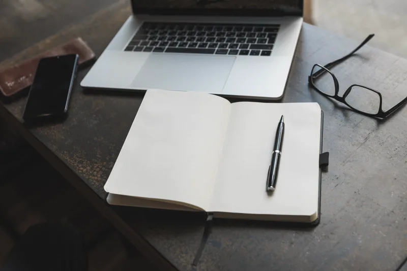 An open notebook with a pen laid across it, beside a laptop and a pair of reading glasses on a worn desk