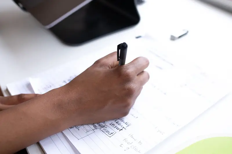 A hand holds a pen over a page of handwritten planning notes beside a laptop